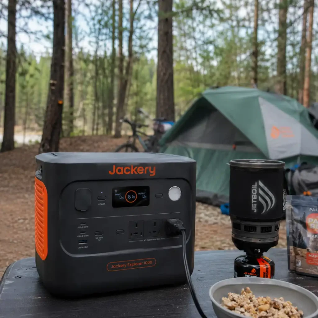 Jackery Explorer 1000 v2 portable power station powering camping equipment at a forest campsite beside a tent.